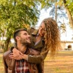 Smiling woman embraces man from behind, piggyback style, in a park setting with autumn foliage. Both are wearing brown jackets.