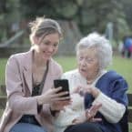Young woman showing something on her smartphone to an elderly woman sitting on a park bench. The older woman is pointing at the screen, seemingly engaged. The scene suggests intergenerational connection and learning.