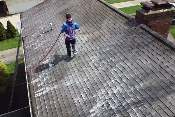 Aerial view of a man power washing a gray shingle roof. The roof cleaning process reveals cleaner shingles, with a chimney and gutter also visible.